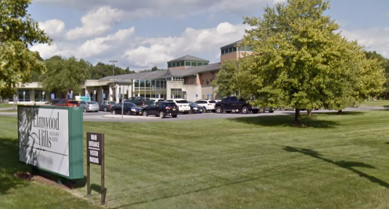 Exterior view of Elmwood Hills Healthcare Center showing the building with large windows, a parking lot with several cars, green grass, and trees under a partly cloudy sky.