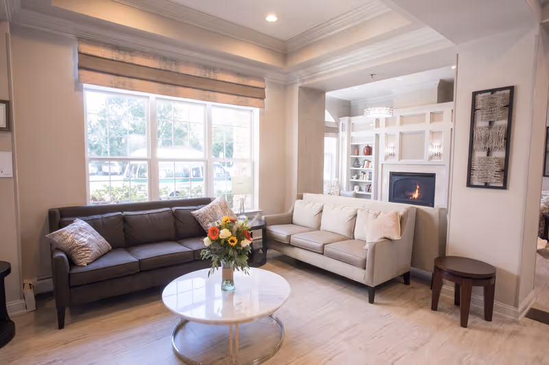 A bright and cozy living room with two sofas, one dark gray and one beige, arranged around a round marble coffee table with a vase of colorful flowers. Large windows with beige Roman shades let in natural light. In the background, there is a fireplace with a fire burning and built-in shelves with decorative items. The walls are light-colored, and there are framed artworks on the walls.