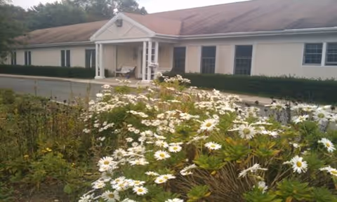 Single-story building with a light-colored exterior and a covered entrance. In the foreground, there is a garden area filled with white daisies and green foliage. The roof appears to have some discoloration or wear.
