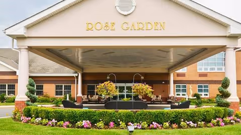 Front entrance of Rose Garden Nursing and Rehabilitation Center featuring a covered drop-off area with seating, surrounded by well-maintained landscaping including green bushes and colorful flowers.