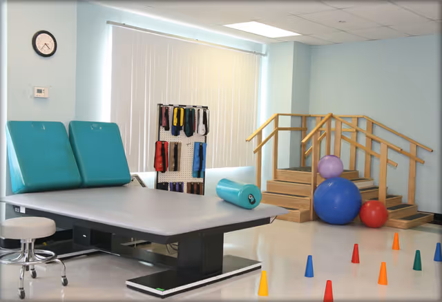 A physical therapy room with a padded treatment table, a stool, colorful exercise bands hanging on a rack, wooden stairs with handrails for rehabilitation, and several exercise balls and small colored cones on the floor.