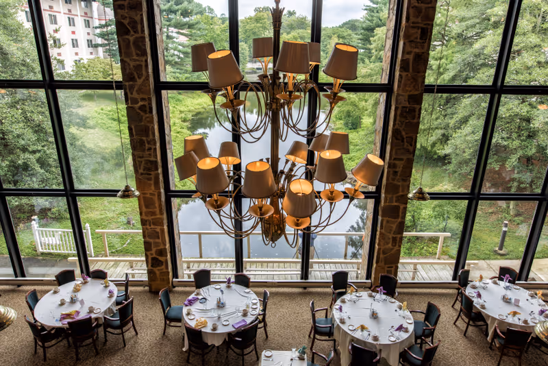 View of a dining room with round tables set with white tablecloths, napkins, cups, and plates. The room features large floor-to-ceiling windows overlooking a pond and greenery outside. A large chandelier with multiple lampshades hangs from the ceiling.