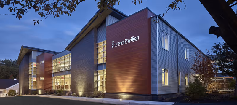 Exterior view of The Shubert Pavilion building at dusk, showing modern architecture with large windows, illuminated from inside, surrounded by trees and landscaping.
