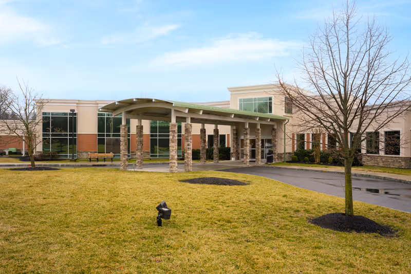 Exterior view of a modern single-story building with large windows and a covered entrance supported by stone pillars. The foreground features a grassy area with a few leafless trees and a paved driveway leading to the entrance under a partly cloudy sky.