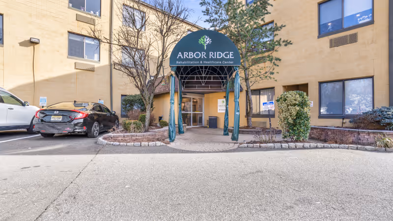 Entrance to Arbor Ridge Rehabilitation & Healthcare Center with a green canopy displaying the facility's name. The building is beige with several windows, and there are a few parked cars and some landscaping with trees and bushes near the entrance.