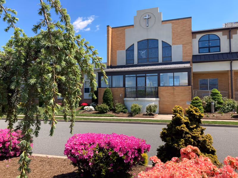 Exterior view of St Joseph's Senior Home - Assisted Living and Nursing Center building with a cross symbol on the upper facade. The foreground features a landscaped garden with vibrant pink and orange flowers, green shrubs, and a tree branch hanging down. The sky is clear and blue.