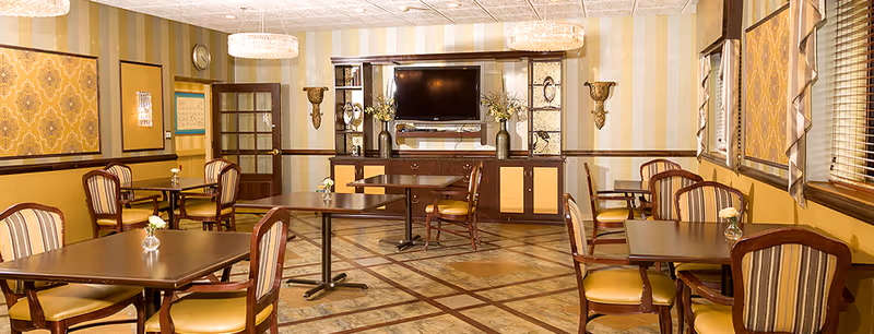 A well-lit common area with several square wooden tables and cushioned chairs arranged neatly. The walls have a striped and patterned wallpaper in shades of yellow and beige. A large flat-screen TV is mounted on a wooden cabinet with decorative vases and flowers. Two chandeliers hang from the ceiling, and windows with blinds and valances are on the right side.