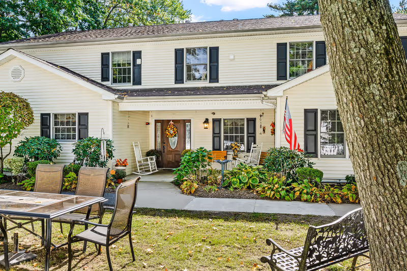 Front exterior view of a two-story white building with black shutters, a wooden front door decorated with a fall wreath, and a covered porch with rocking chairs and a bench. There are green shrubs and plants along the walkway leading to the entrance. An American flag is displayed near the porch. In the foreground, there is a patio area with a glass-top table and several chairs, as well as a decorative metal bench and a large tree trunk.