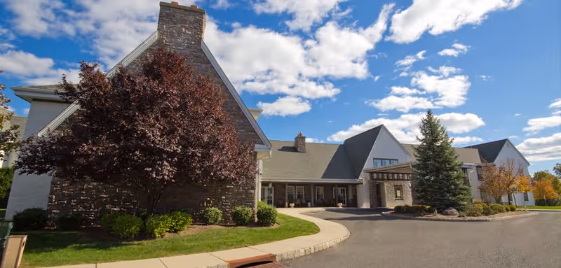 Exterior view of Independence Manor Hunterdon, showing a large building with stone and white siding, a steep roof, and a covered entrance. The foreground features a curved driveway, manicured lawn, and several trees under a partly cloudy blue sky.