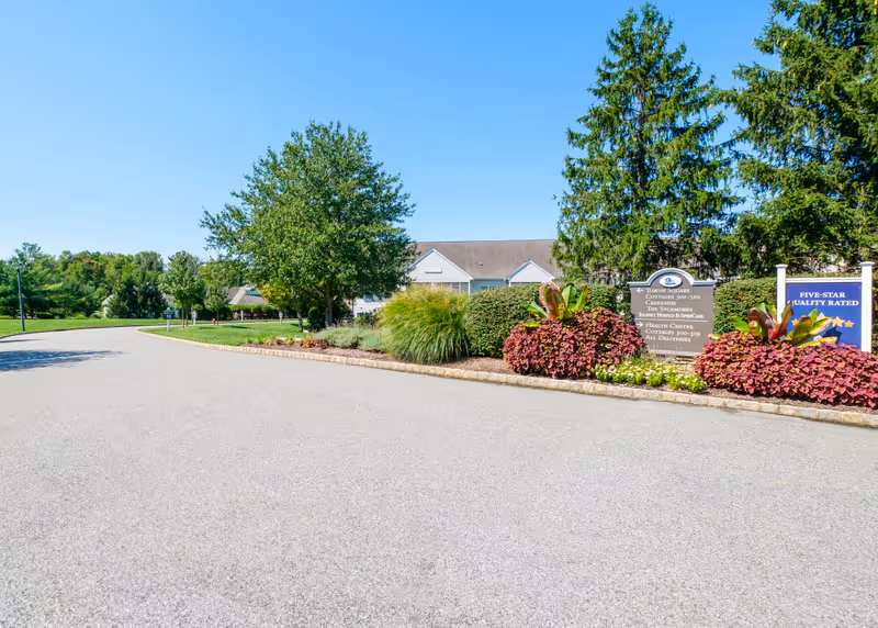 A paved road curves gently to the left, bordered by well-maintained landscaping with bushes, flowers, and trees. In the background, there are residential buildings with light-colored siding and sloped roofs. Two signs are visible on the right side of the road, one indicating directions to Towne Square, cottages, health center, and other facilities, and the other sign displaying a five-star quality rating.