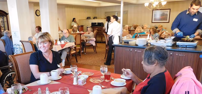 Seniors and staff eating and socializing at tables in a communal dining room with a food service counter.