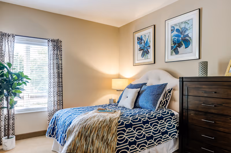 A cozy bedroom with a bed featuring a blue and white patterned bedspread and multiple pillows. There is a wooden dresser with decorative items on top, two framed floral artworks on the beige wall above the bed, a window with patterned curtains, and a potted plant beside the window. A bedside lamp emits warm light.