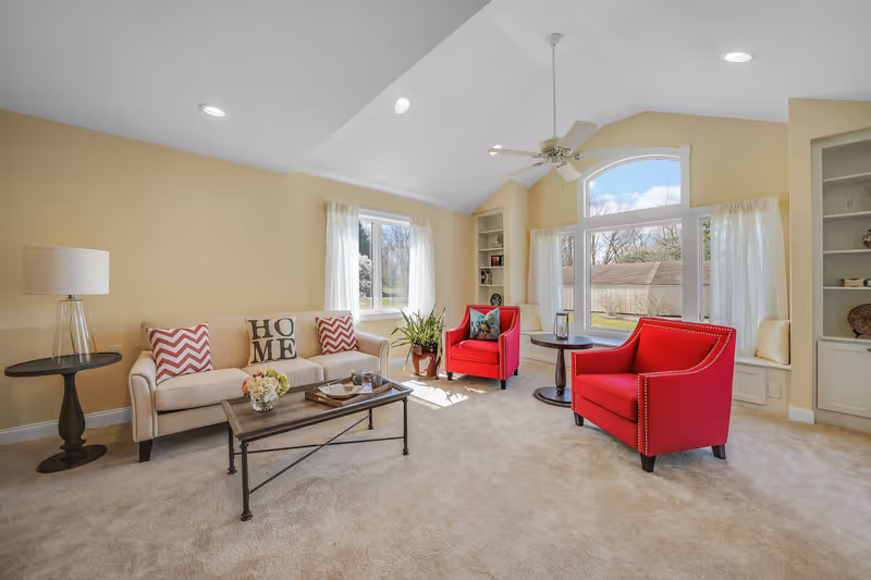A bright living room with beige walls and carpet, featuring a cream-colored sofa with decorative pillows, two red armchairs, a wooden coffee table with a flower arrangement, a side table with a lamp, built-in shelves, large windows with white curtains, and a ceiling fan.
