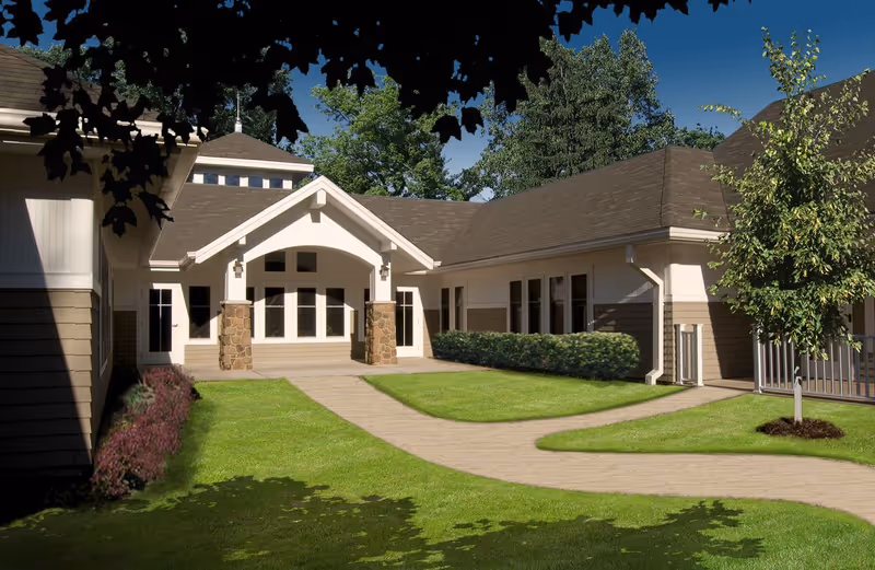 Entrance courtyard and front façade of a single-story senior living building with a covered entry, walkway, lawn, and landscaping.