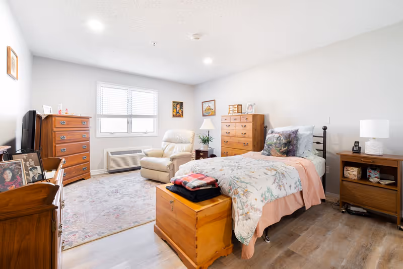 A bright and cozy bedroom in an assisted living facility featuring a single bed with floral bedding and a decorative pillow, a wooden chest at the foot of the bed with folded blankets on top, a beige recliner chair near a window with white blinds, two wooden dressers, a nightstand with a lamp and phone, framed pictures on the walls, and a patterned area rug on the wooden floor.