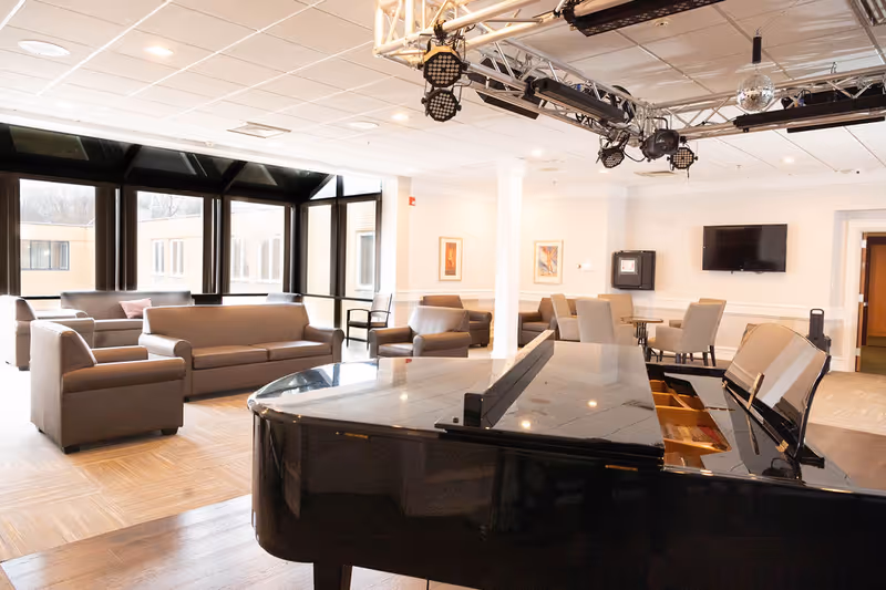 A spacious senior living common area with a glossy black grand piano in the foreground, several brown sofas and armchairs arranged near large windows letting in natural light, and a wall-mounted TV with framed artwork on the walls.