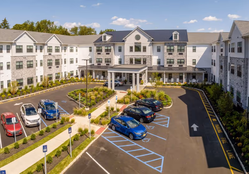 Front exterior view of Azalea at Cinnaminson senior living facility showing a three-story building with white and stone facade, a covered entrance, landscaped greenery, and a parking lot with several cars and handicap parking spaces.