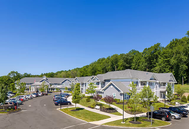 Exterior view of Brightview Warren - Senior Assisted Living & Memory Care facility showing a large, two-story building with gray siding and white trim surrounded by landscaped greenery and a parking lot with multiple cars under a clear blue sky.