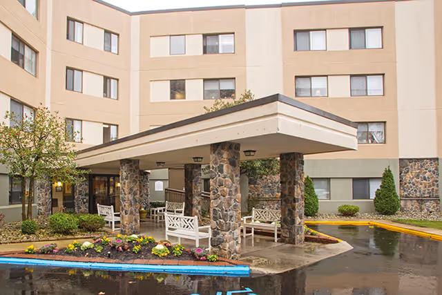 Exterior view of a multi-story senior living facility with a covered entrance supported by stone pillars. There are white benches under the entrance canopy, landscaped flower beds, small trees, and shrubs around the driveway area.