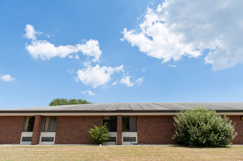 Exterior view of a single-story brick building with a flat roof under a blue sky with scattered clouds. There are a few small bushes in front of the building and a grassy area in the foreground.