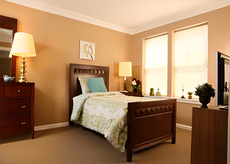 Warm, neatly arranged bedroom with a wooden bed, bedside tables and lamps, a dresser, and two windows with shades.