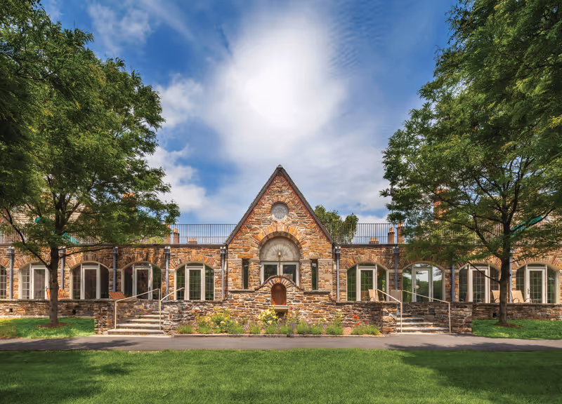Exterior view of a stone building with a triangular central section and multiple arched windows. The building is surrounded by green grass and trees under a partly cloudy blue sky.