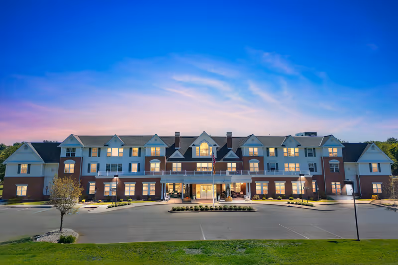 Front exterior view of Brighton Gardens of West Orange, a large multi-story senior living facility with a brick and white facade, multiple windows lit from inside, a central entrance with a covered porch, an American flag in front, and a parking area with some landscaping under a clear blue and pink sky at dusk.