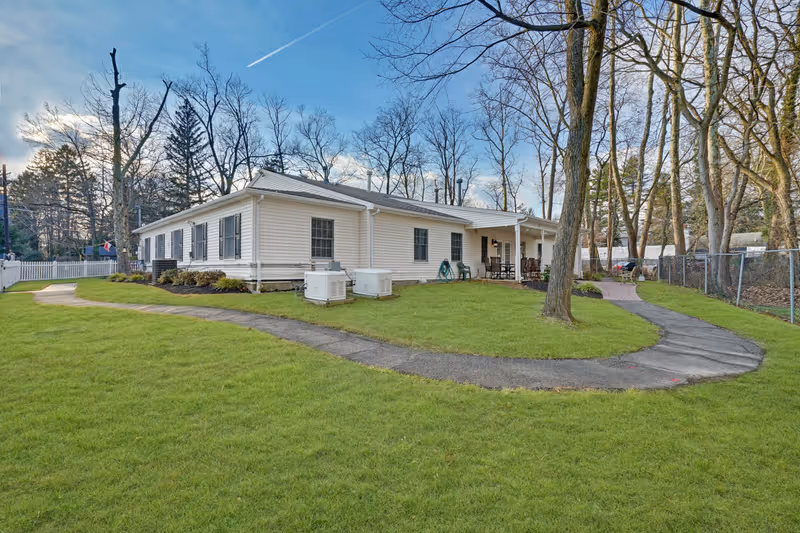 Exterior view of a single-story white building with black shutters surrounded by a green lawn and trees. A paved walkway curves around the building, and there is a covered patio area with outdoor furniture. The sky is partly cloudy with a visible contrail.