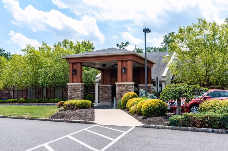 Entrance to a senior living facility with a covered drop-off area supported by stone and wood pillars, surrounded by well-maintained landscaping including bushes and trees, with a red car parked nearby under a bright blue sky with some clouds.