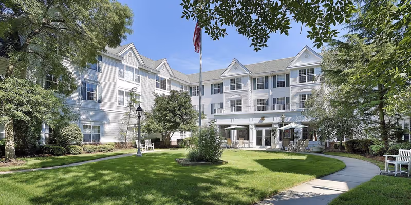 A bright and sunny courtyard area of a senior living facility with a well-maintained lawn, trees, and shrubs. The three-story white building with multiple windows surrounds the courtyard. There are benches and tables with umbrellas for outdoor seating, and an American flag is flying on a flagpole in the center.