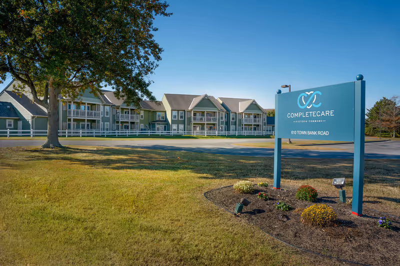 Exterior view of Complete Care at Victoria Commons facility with a large sign displaying the name and address 610 Town Bank Road. The building is two stories with balconies and surrounded by a white fence, trees, and a grassy area under a clear blue sky.