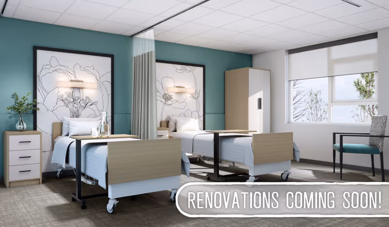 A bright and modern shared bedroom in a healthcare facility with two hospital beds separated by a privacy curtain. Each bed has a bedside table with a vase and flowers, and large floral artwork on the teal-colored wall behind the beds. There is a window with a roller shade letting in natural light, a chair, and a wardrobe in the room. Text overlay at the bottom right reads 'RENOVATIONS COMING SOON!'