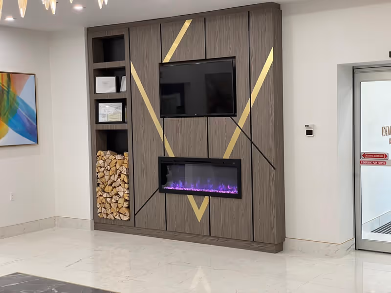 A modern interior wall with a mounted flat-screen TV above an electric fireplace with purple flames. The wall has a wood panel design with gold diagonal accents. To the left of the TV and fireplace is a built-in shelving unit containing stacked firewood and framed certificates. A colorful abstract painting is hung on the adjacent wall. To the right is a glass door with signage and a thermostat on the wall.