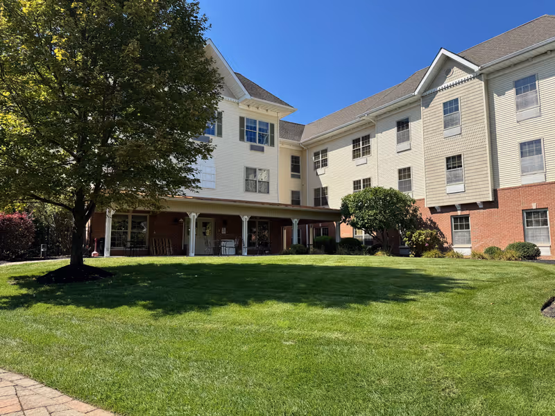Front exterior of a multi-story assisted living building with a green lawn, tree, and covered patio under a blue sky.