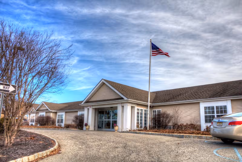 Exterior view of Merion Gardens Assisted Living facility showing a single-story building with beige siding, a peaked roof, and a covered entrance. An American flag is flying on a flagpole near the entrance. There is a car parked in a handicapped parking space to the right, and leafless trees and shrubs surround the building under a partly cloudy sky.