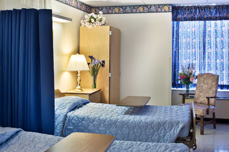 A senior living facility bedroom with two hospital-style beds covered in blue quilted bedspreads. Each bed has an overbed table. A wooden nightstand with a lamp and a vase of flowers is positioned between the beds. There is a wooden wardrobe in the corner with a floral arrangement on top. A floral-patterned armchair sits near a window with blue floral curtains, and a small table next to it holds another vase of flowers. A blue privacy curtain is partially visible on the left side of the image.