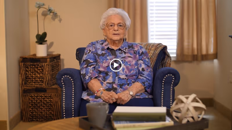 An elderly woman with white hair and glasses sits in a blue armchair in a cozy room. She is wearing a purple and blue patterned blouse and has bracelets on her wrist. Behind her, there is a window with beige curtains and a small table with a potted orchid plant. In front of her is a coffee table with a tray holding a cup, books, and a decorative spherical object.