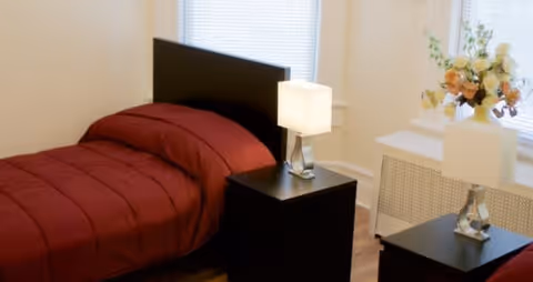 A bedroom with a single bed covered in a red comforter, a black headboard, and two black nightstands each with a modern lamp. There is a window with blinds behind the bed and a white radiator cover with a vase of flowers on top.