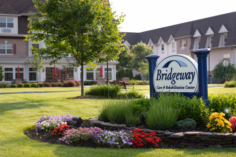 Landscaped lawn and flowerbed with a prominent 'Bridgeway Care & Rehabilitation Center' sign in front of a multi-story senior living building.