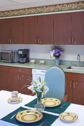 A kitchen area with wooden cabinets, a microwave, a coffee maker, and a sink. In the foreground, a dining table is set with plates, cups, and silverware on green placemats, with a vase of white flowers as a centerpiece. Another vase with purple and white flowers is placed on the countertop.