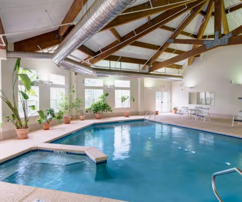 Indoor swimming pool area with clear blue water, surrounded by potted plants and several windows letting in natural light. The ceiling features exposed wooden beams and large metal air ducts. There are a few tables and chairs along the far wall.