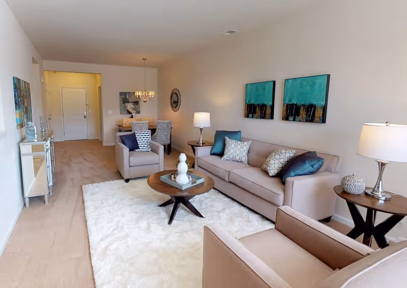 Bright furnished living room with beige sofas and armchairs, a round coffee table on a white rug, and a dining area visible in the background.