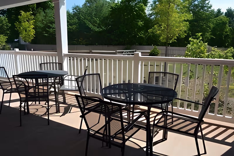 Outdoor patio area with several black metal tables and chairs on a covered deck. The deck has a white railing and overlooks a green area with trees and bushes under a clear blue sky.