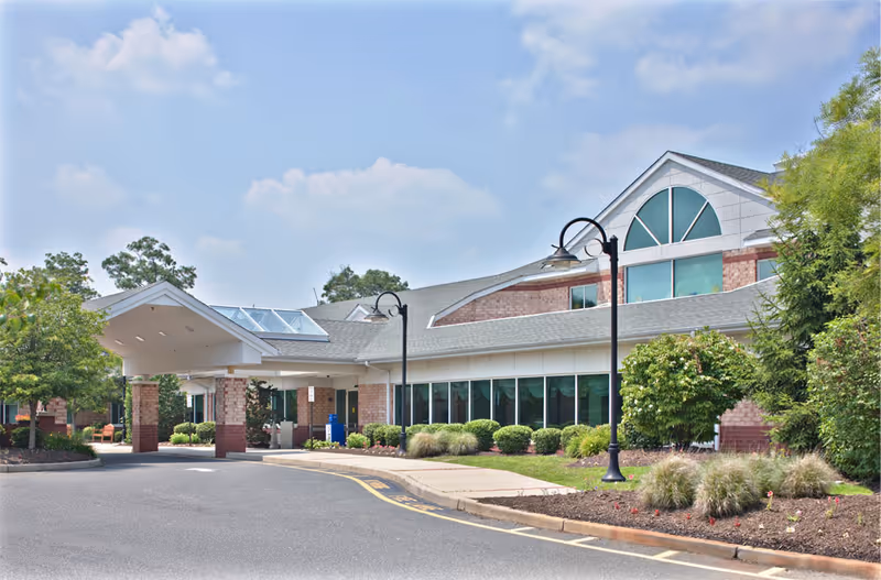 Exterior view of a single-story brick building with large windows and a covered entrance. The building is surrounded by landscaped bushes, trees, and a paved driveway under a partly cloudy sky.