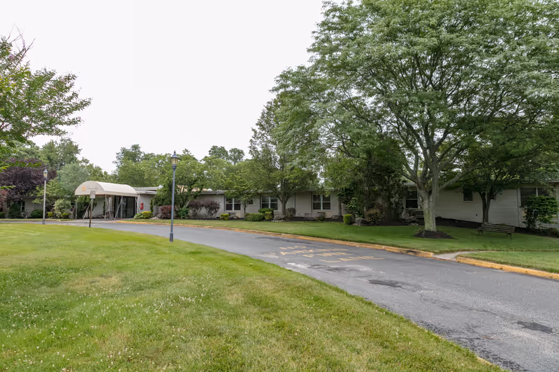 Exterior view of Tower Lodge Care Center showing a single-story building with a covered entrance, surrounded by green trees and grass, with a paved driveway leading to the entrance.