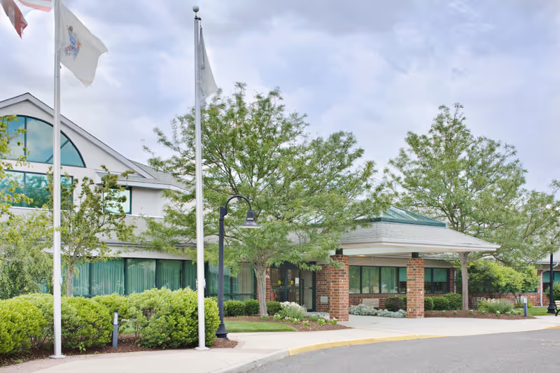 Exterior view of Southern Ocean Center showing the entrance with a covered drop-off area, brick pillars, large windows, and surrounding greenery including trees and bushes. Two flagpoles with flags are visible in front of the building under a cloudy sky.