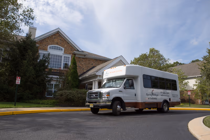 A white shuttle bus parked in front of a brick and stone senior living facility building with trees and shrubs around. The bus has signage for Spring Village at Galloway and Senior Living services.