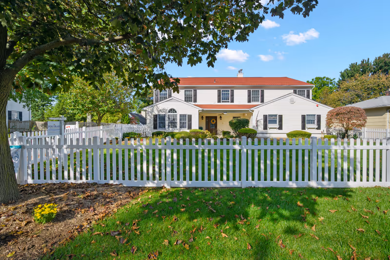 Exterior view of a two-story white building with black shutters and a red roof, surrounded by a white picket fence and green lawn with trees and shrubs under a clear blue sky.