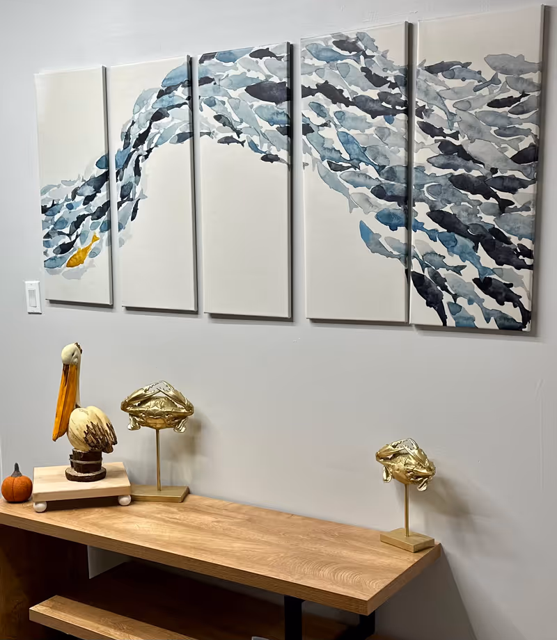 A wooden console table against a light gray wall with decorative items including a wooden pelican sculpture, two gold crab figurines on stands, and a small pumpkin. Above the table is a six-panel wall art piece depicting a school of fish swimming, mostly in shades of blue and gray with one yellow fish.