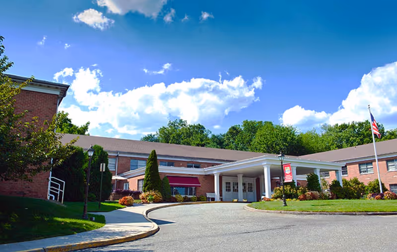 Exterior view of Emerson Health and Rehabilitation Center, a single-story brick building with a covered entrance, surrounded by greenery and a clear blue sky with some clouds.